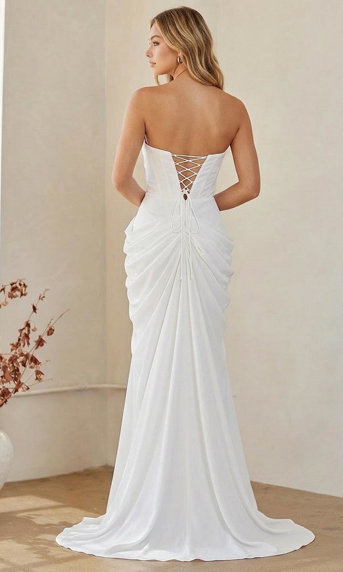 A woman faces away in a neutral-toned room, showcasing the Long Formal Dress: Amelia Couture 6149—a strapless gown with a lace-up back and gathered draping.